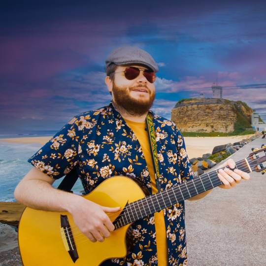 Man in a cap on a beach playing a guitar wearing sunglasses in a floral shirt with a sunset in the background